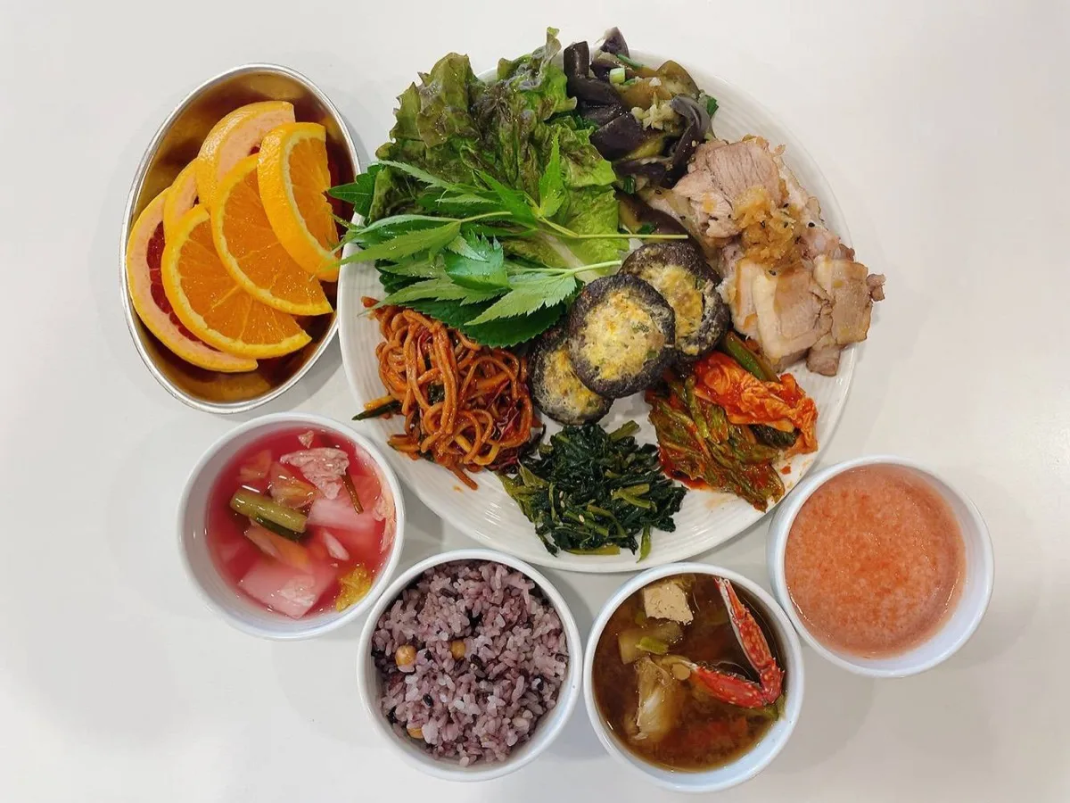 식단 구성 — 흑미 잡곡밥, 수육, 상추·깻잎 쌈채소, 표고버섯전, 잡채, 시금치나물, 배추김치, 게 미역국, 과일 주스, 물김치, 오렌지·자몽